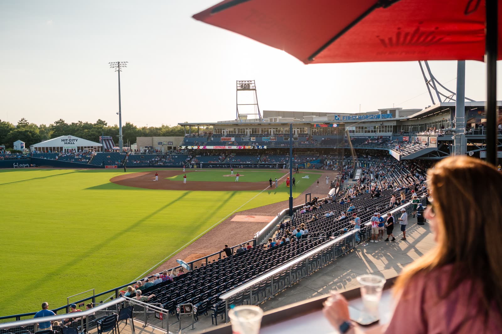 A ballpark at golden hour seen from a private suite, a red umbrella overhead