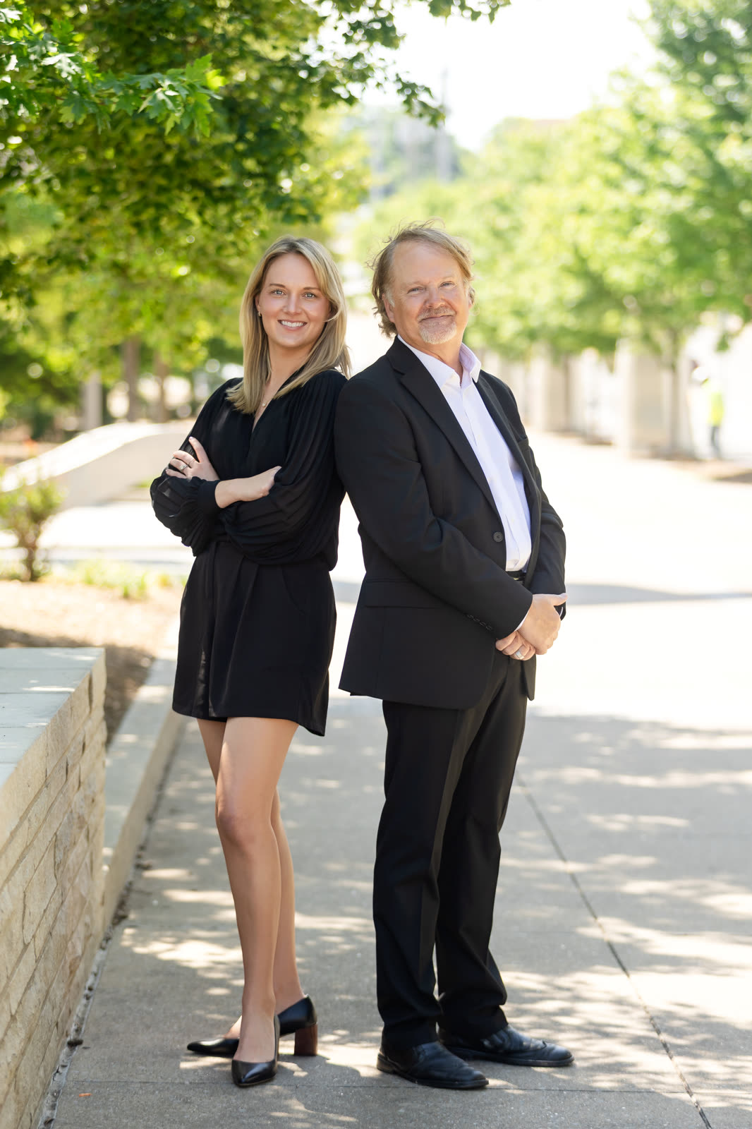 Two colleagues back to back on a sunny tree-lined walkway, her in black, him in a suit