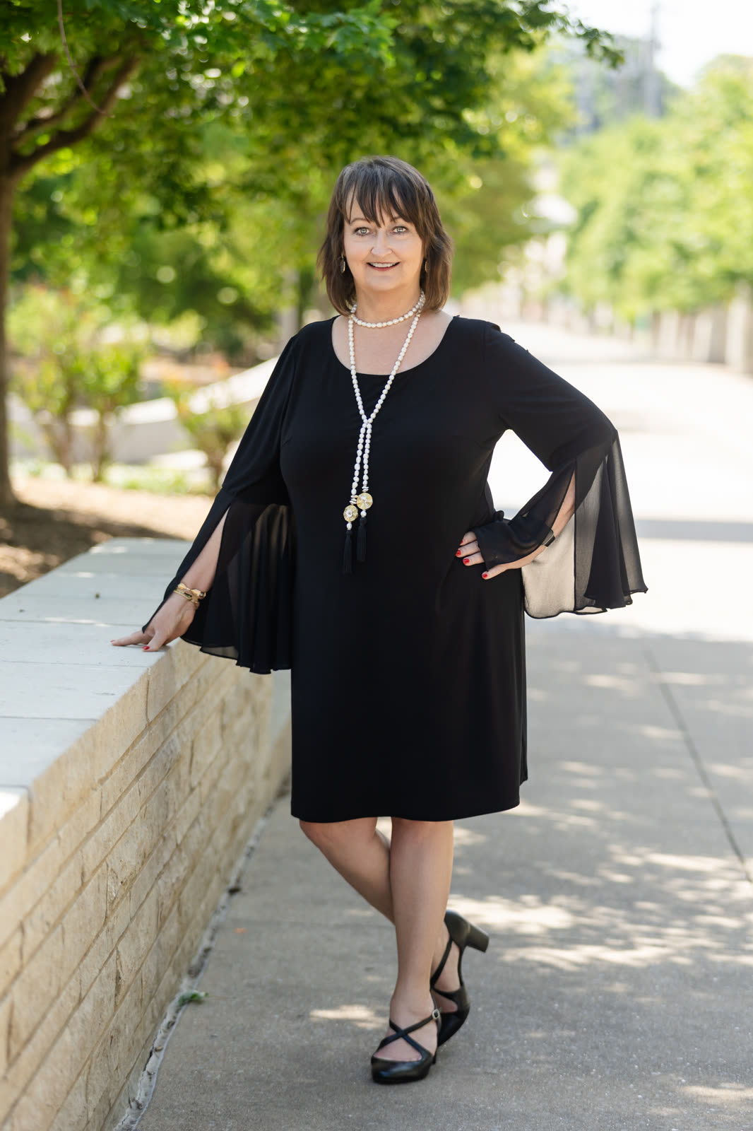A lifestyle headshot of a woman in black beside a stone wall on a tree-lined walkway