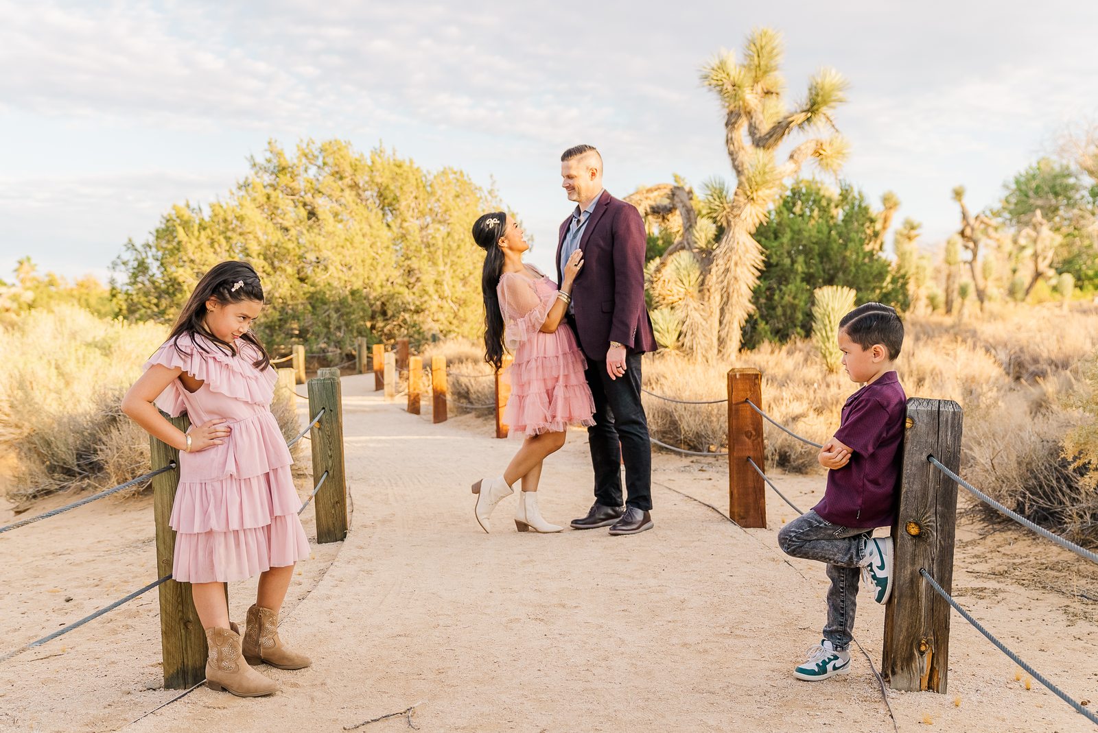 A family of four on a desert path at golden hour, the parents dancing while their two children lean against fence posts