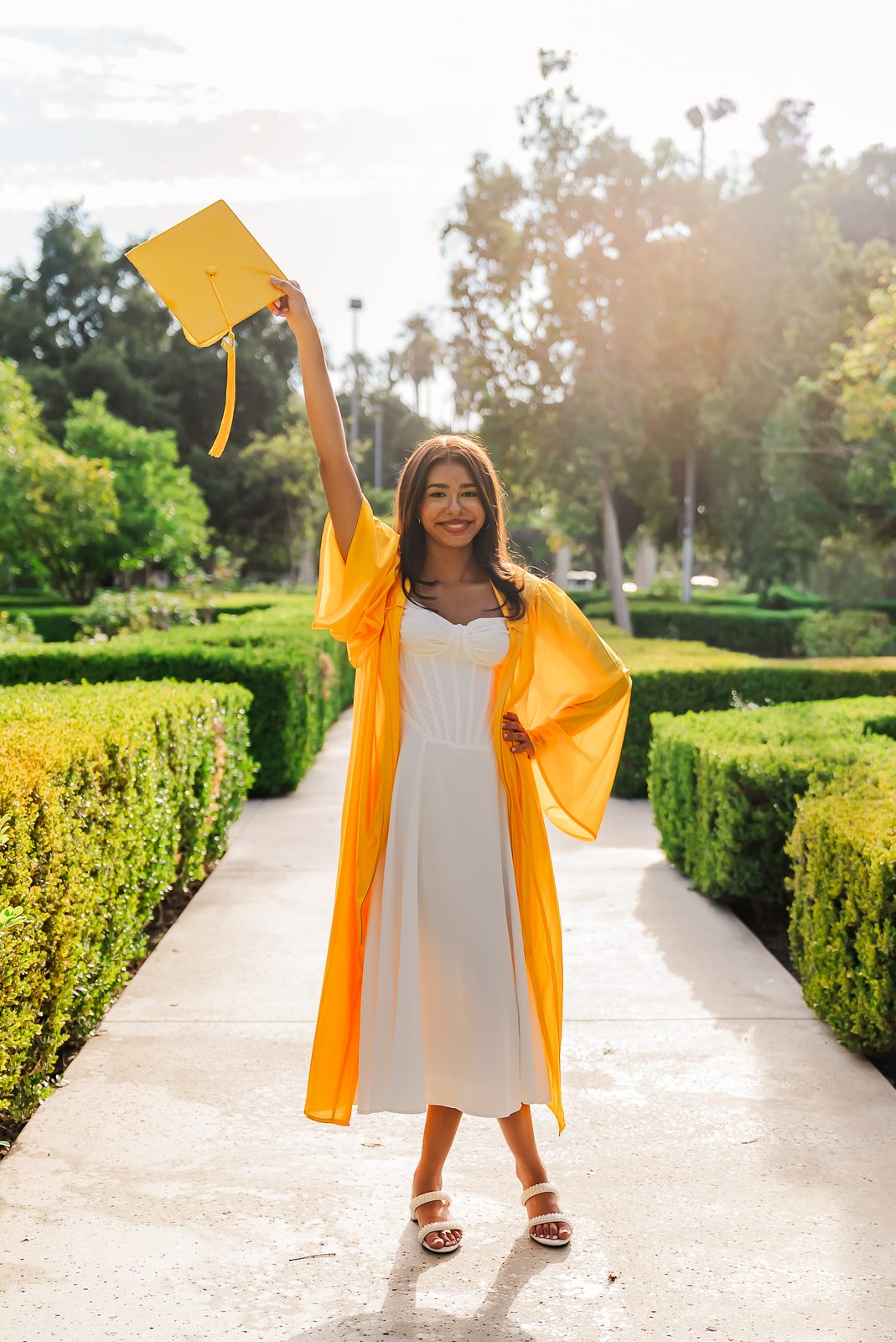 A graduate holding her cap overhead, backlit by late sun