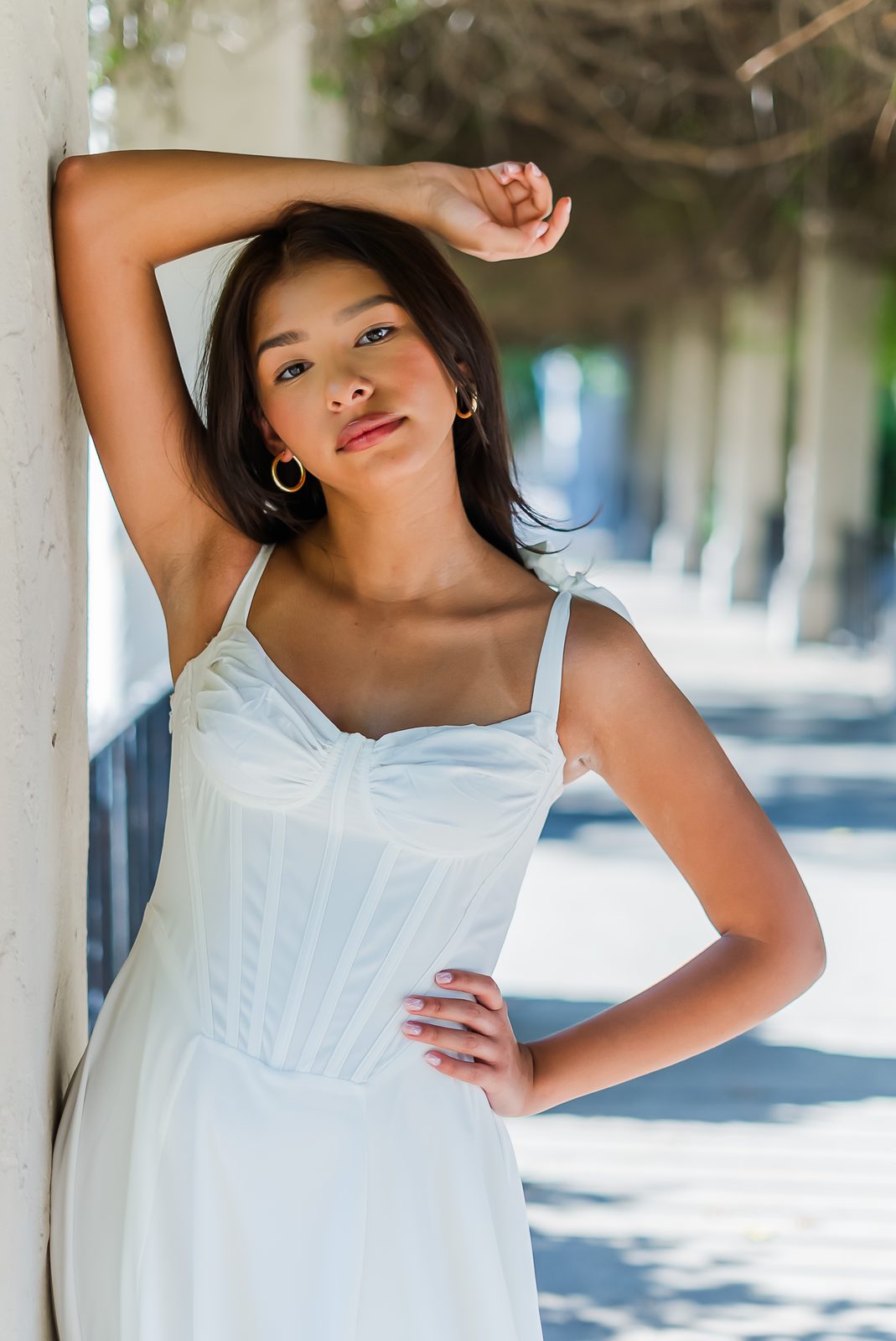 A senior in a white dress with one arm raised against a shaded colonnade