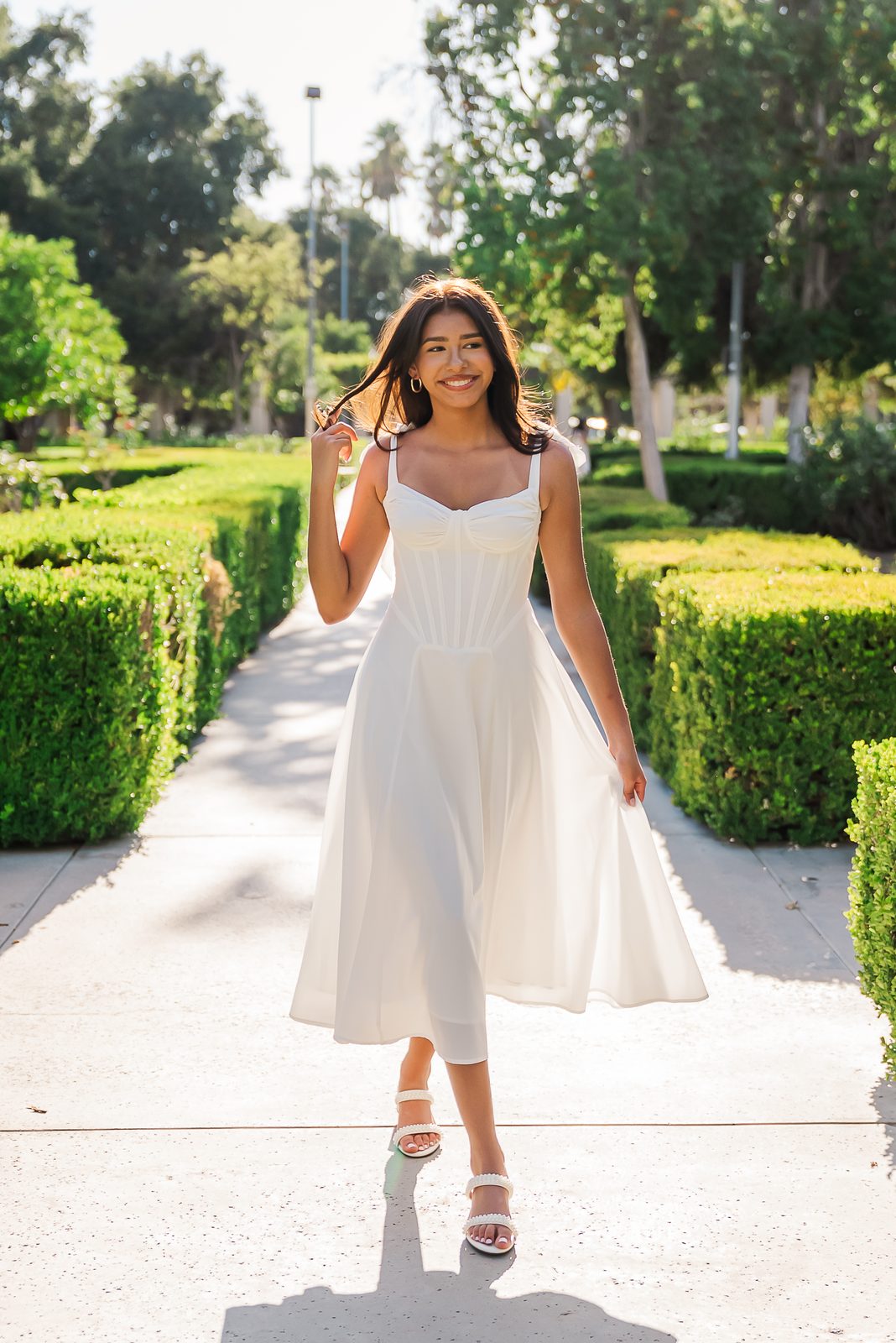 A senior walking toward camera on a garden path in a white dress