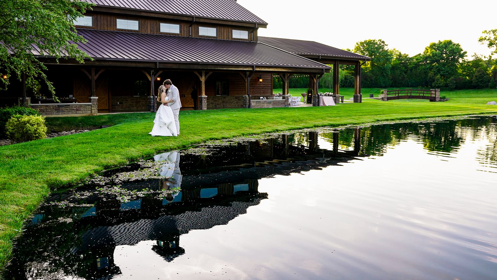 A couple kissing beside a pond, their reflection in the water and a timber barn venue behind