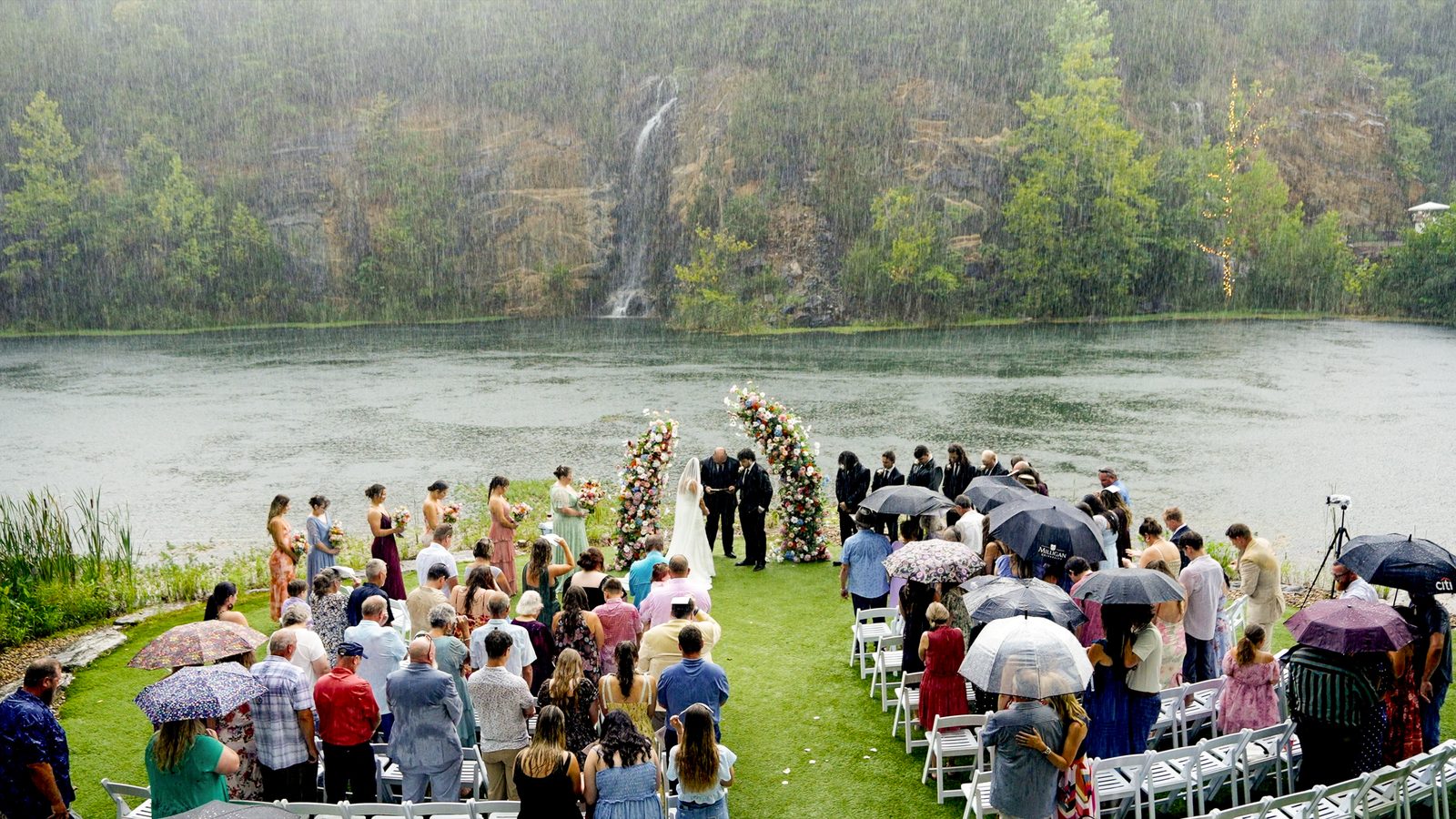 A couple exchanging vows in pouring rain beside a quarry lake and waterfall, guests standing under umbrellas