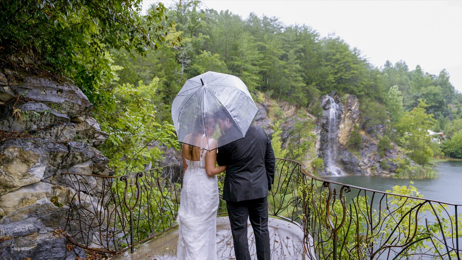 A bride and groom touching foreheads under a clear umbrella in the rain, a waterfall behind them