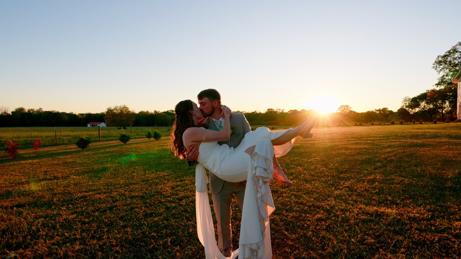 A groom lifting his bride mid-kiss as the sun sets behind an open field